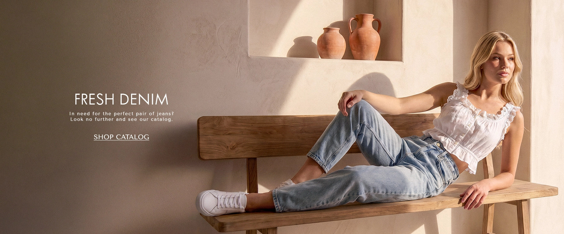 Woman sitting on a wooden bench wearing light blue jeans and a white top, with a shelf of vases in the background.